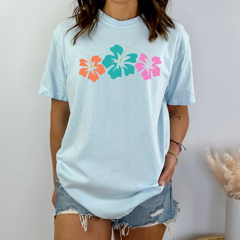 May include: A light blue t-shirt with a row of three colorful hibiscus flowers printed on the front. The flowers are pink, orange, and teal.
