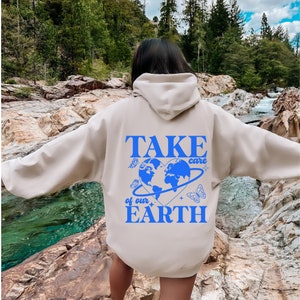 Op de afbeelding: Een beige hoodie met een blauwe afbeelding op de rug die "TAKE care of our EARTH" zegt met een blauwe hartvormige wereld en vlinders.
