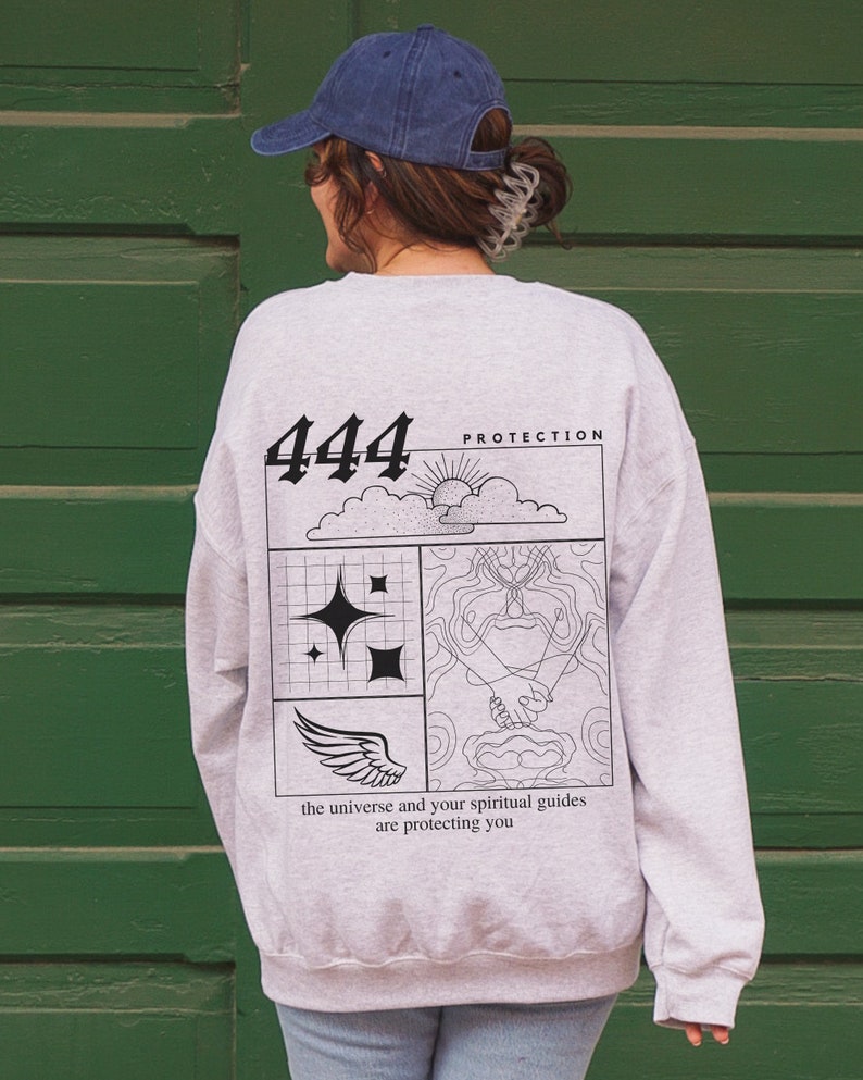 May include: A gray sweatshirt with a black graphic design on the back. The graphic features the number 444, a sun, clouds, stars, a hand holding a hand, and wings. The text "Protection" and "the universe and your spiritual guides are protecting you" are also included in the design.