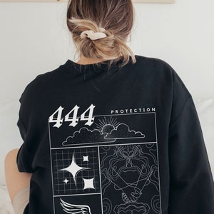 May include: Black sweatshirt with a white graphic design featuring the number 444, the words "Protection", a sun, clouds, stars, a wing, and two hands clasped together. The text below the graphic reads "the universe and your spiritual guides are protecting you".