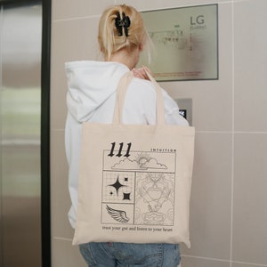 K&ouml;nnte beinhalten: Eine beige Tragetasche mit schwarzem Text und Grafiken. Der Text lautet "111 Intuition" und "trust your gut and listen to your heart".