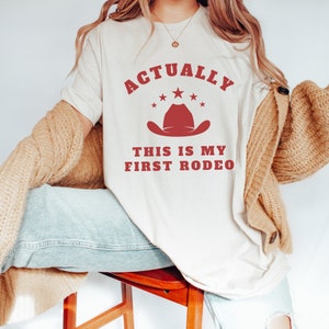 K&ouml;nnte beinhalten: Ein wei&szlig;es T-Shirt mit dem Text "Actually this is my first rodeo" in Rot. Ein roter Cowboyhut mit Sternen befindet sich &uuml;ber dem Text.