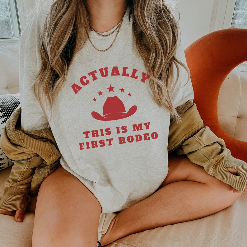 K&ouml;nnte beinhalten: Ein graues Sweatshirt mit dem Text "Actually This Is My First Rodeo" in roten Buchstaben. &Uuml;ber dem Text befindet sich ein roter Cowboyhut mit f&uuml;nf wei&szlig;en Sternen.