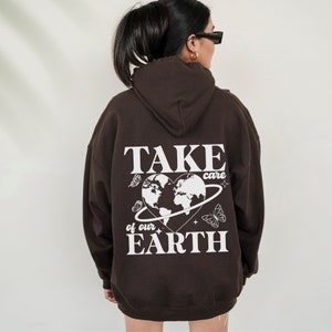 Op de afbeelding: Bruin hoodie met een witte grafische print op de rug. De print toont een hartvormige wereld met de tekst "TAKE care of our EARTH" en vlinders.