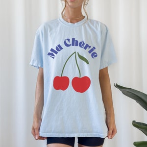 May include: A light blue t-shirt with a graphic of two red cherries and the text "Ma Ch&eacute;rie" in blue.