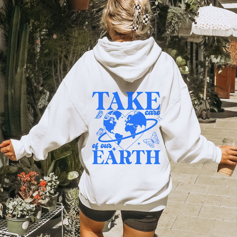 Op de afbeelding: Witte hoodie met een blauwe afbeelding met de tekst "TAKE care of our EARTH" met een blauwe hartvormige wereld en vlinders.
