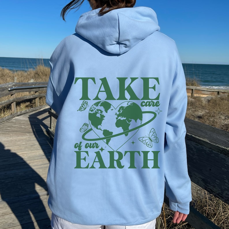 Op de afbeelding: Een lichtblauwe hoodie met een groene afbeelding op de rug die "TAKE care of our EARTH" zegt met een groene hartvormige wereld en vlinders.