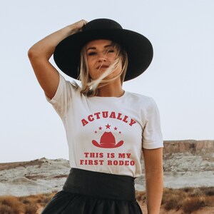 K&ouml;nnte beinhalten: Wei&szlig;es T-Shirt mit rotem Text, der "Actually This Is My First Rodeo" lautet, und einem roten Cowboyhut-Motiv. Das Shirt wird von einer Person getragen, die einen schwarzen Hut und einen schwarzen Rock tr&auml;gt.