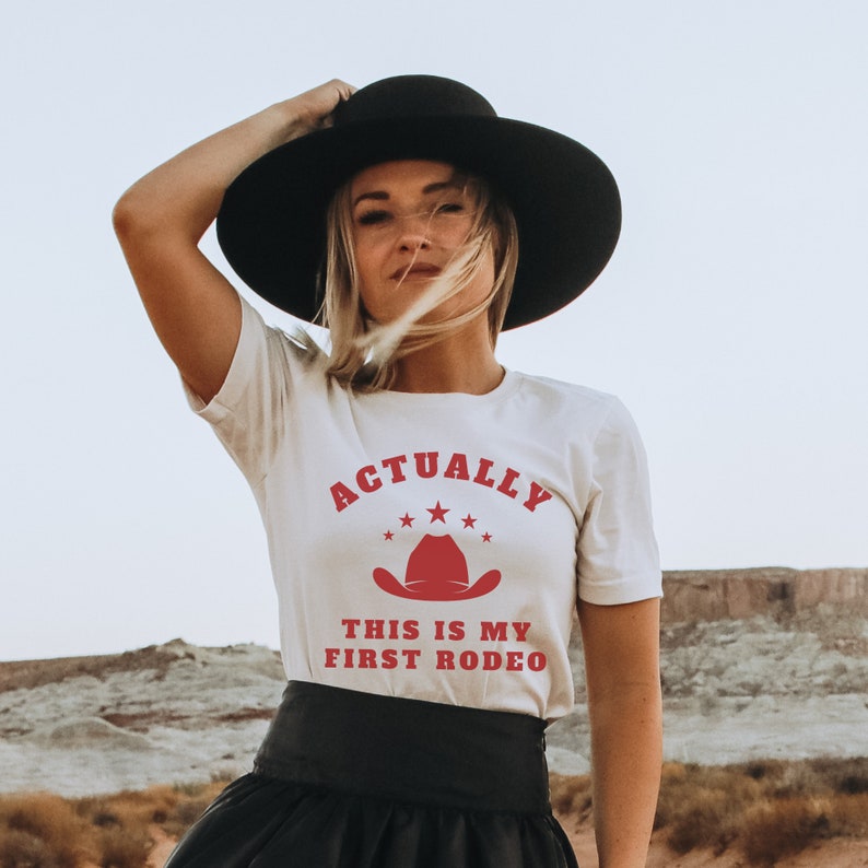 K&ouml;nnte beinhalten: Wei&szlig;es T-Shirt mit rotem Text, der "Actually This Is My First Rodeo" lautet, und einem roten Cowboyhut-Motiv. Das Shirt wird von einer Person getragen, die einen schwarzen Hut und einen schwarzen Rock tr&auml;gt.