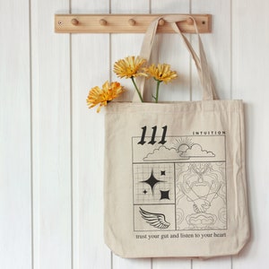 K&ouml;nnte beinhalten: Eine beige Tragetasche mit schwarzem Text und Grafiken. Der Text lautet "111 Intuition" und "vertraue deinem Bauchgef&uuml;hl und h&ouml;re auf dein Herz". Die Grafiken beinhalten eine Sonne, Wolken, Sterne und eine Hand, die ein Herz h&auml;lt.