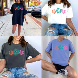 May include: Three different color variations of a short-sleeved t-shirt with a graphic of three hibiscus flowers. The shirts are shown on models wearing distressed denim shorts.