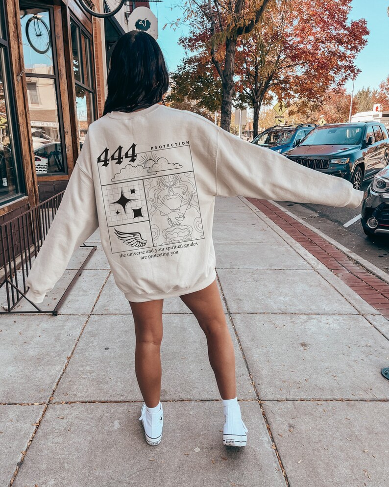 May include: A beige sweatshirt with a graphic print on the back. The graphic features the number 444, a sun, clouds, a wing, a hand, and the text "Protection, the universe and your spiritual guides are protecting you".
