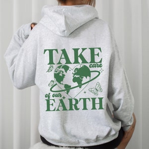 Op de afbeelding: Een grijze hoodie met een groene afbeelding op de rug. De afbeelding toont een hartvormige aarde met een ring eromheen, vlinders en de tekst "TAKE care of our EARTH".