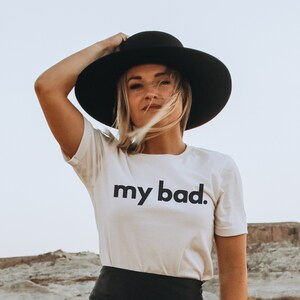May include: A woman wearing a black wide-brimmed hat and a cream-colored t-shirt with the words "my bad" printed in black. The background shows a desert landscape.