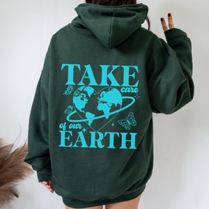 Op de afbeelding: Een donkergroene hoodie met een blauwe afbeelding op de rug die "TAKE care of our EARTH" zegt met een hartvormige wereld en vlinders.