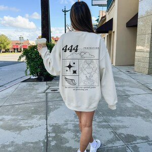 May include: A cream-colored sweatshirt with a black graphic design on the back. The graphic features the number 444, the words "Protection" and "the universe and your spiritual guides are protecting you", and a stylized design of a sun, clouds, stars, hands, and wings.