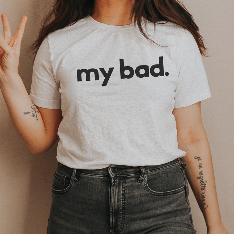 May include: A light gray t-shirt with the phrase "my bad." printed in bold black letters. The shirt is paired with dark gray jeans. The image shows a person wearing the shirt.