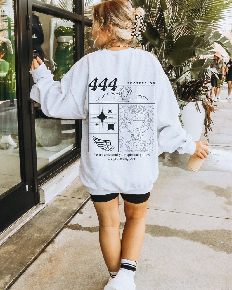 May include: A gray sweatshirt with a graphic design on the back. The design features a black and white illustration of a cloud, sun, stars, wings, and hands. The text "444 Protection" and "the universe and your spiritual guides are protecting you" is printed on the sweatshirt.
