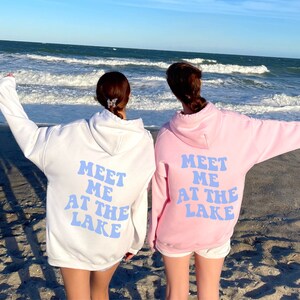 May include: Two people wearing matching hoodies with the text "Meet me at the lake" in blue letters on the back. The hoodies are white and pink. The people are standing on a sandy beach with the ocean in the background.