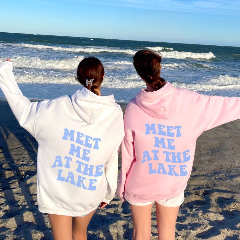 May include: Two people wearing matching hoodies with the text "Meet me at the lake" in blue letters on the back. The hoodies are white and pink. The people are standing on a sandy beach with the ocean in the background.