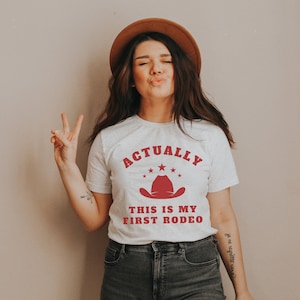 K&ouml;nnte beinhalten: Ein wei&szlig;es T-Shirt mit dem Text "Actually This Is My First Rodeo" in roten Buchstaben. &Uuml;ber dem Text befindet sich ein roter Cowboyhut mit f&uuml;nf roten Sternen.