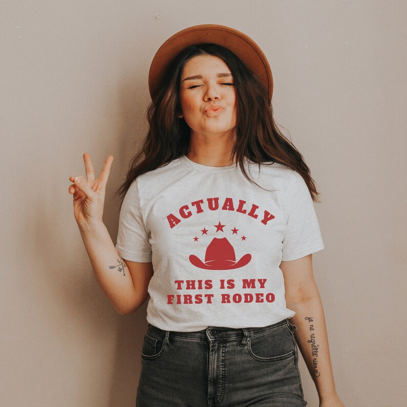 K&ouml;nnte beinhalten: Ein wei&szlig;es T-Shirt mit dem Text "Actually This Is My First Rodeo" in roten Buchstaben. &Uuml;ber dem Text befindet sich ein roter Cowboyhut mit f&uuml;nf roten Sternen.