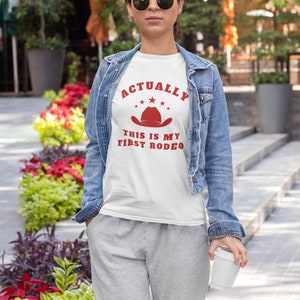 K&ouml;nnte beinhalten: Wei&szlig;es T-Shirt mit rotem Text, der "Actually This Is My First Rodeo" lautet, und einem roten Cowboyhut mit Sternen dar&uuml;ber.