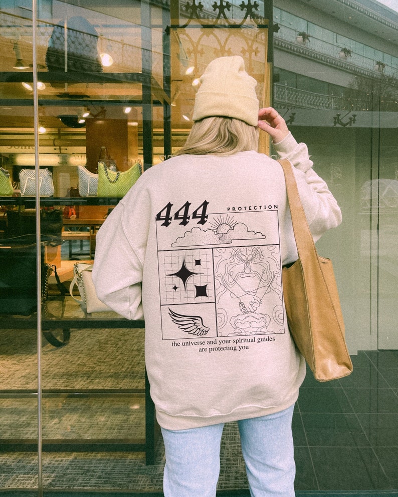 May include: A beige sweatshirt with a black graphic on the back. The graphic features the number 444, a cloud, a sun, a wing, and a hand. The text "Protection" and "the universe and your spiritual guides are protecting you" are also included in the graphic.