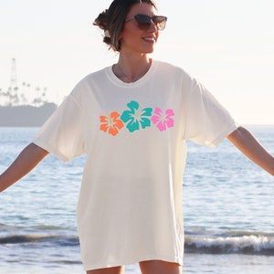 May include: A white t-shirt with a row of three colorful hibiscus flowers printed on the front. The flowers are orange, turquoise, and pink.