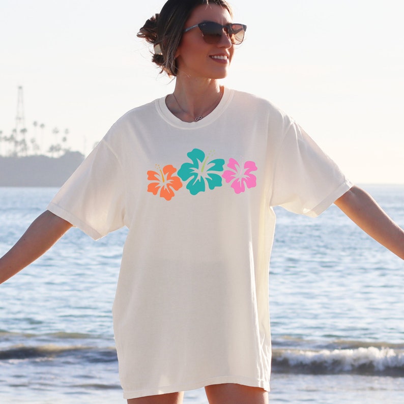 May include: A white t-shirt with a row of three colorful hibiscus flowers printed on the front. The flowers are orange, turquoise, and pink.