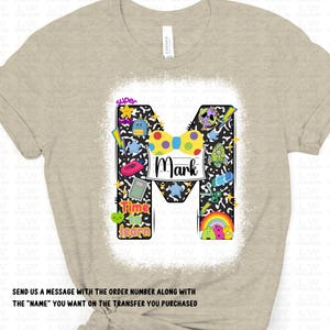 May include: Beige t-shirt with a large, decorated letter "M" featuring a colorful bow tie and the name "Mark". The letter is filled with various cartoon graphics and the words "Super" and "Time to Learn".