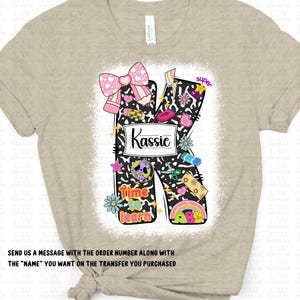 May include: Beige t-shirt featuring a large, decorated letter "K" with the name "Kassie". The letter is filled with colorful school-themed graphics, a pink bow, and the words "Time to learn". The shirt has a distressed, bleached effect.