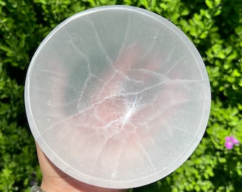 May include: A round, white selenite bowl with a cracked, textured surface. The bowl is held in a hand, with a green leafy background. The bowl is a light, translucent color.