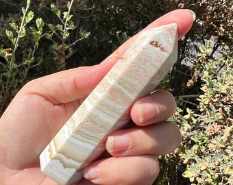 May include: A white and beige banded crystal point, also known as a wand, with a smooth, polished finish. The crystal is held in a hand, showcasing its size and shape.