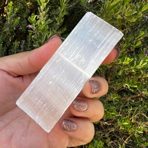 May include: A white selenite crystal slab with a smooth, striated surface. The crystal is held in a hand with glitter nail polish.
