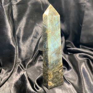 May include: A dark gray labradorite crystal point with iridescent blue and green flashes. The crystal is polished and has a pointed tip.
