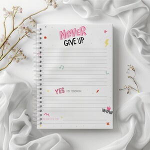 Op de afbeelding: Een wit spiraalnotitieblok met een motiverende quote "Never Give Up" in roze letters. Het notitieblok heeft verschillende kleurrijke illustraties en tekst, waaronder "Yes for tomorrow", "I'm grateful for" en "Wow".