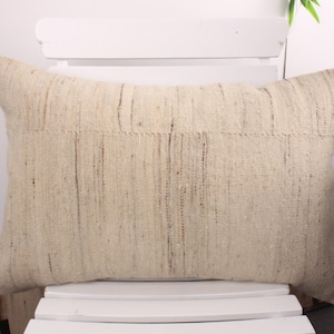 May include: A rectangular beige pillow with a textured surface and dark brown vertical lines. The pillow is placed on a white chair. The pillow has a neutral color palette and a rustic aesthetic, suitable for home decor.