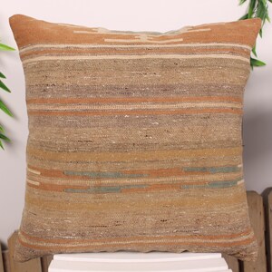 May include: Square decorative pillow with a woven, striped pattern in shades of tan, brown, and orange. The pillow has a textured appearance and is likely made of natural fibers. The design features horizontal stripes and a geometric pattern at the top.