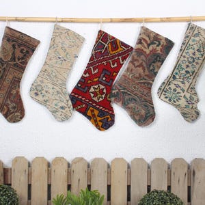 Può includere: Cinque calze di Natale colorate realizzate con tappeti vintage sono appese a un'asta di legno. Le calze sono decorate con motivi intricati in tonalità di rosso, marrone, beige e blu.