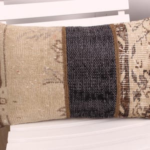 May include: Rectangular decorative pillow with a patchwork design. The pillow features a central dark gray section flanked by beige and patterned sections with brown accents. The pillow is placed on a white chair.