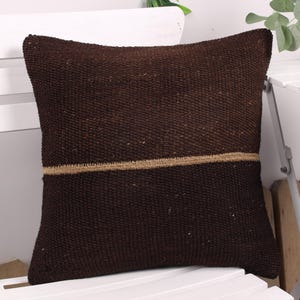 Peut inclure: Un coussin carré marron foncé avec une texture tissée. Une bande horizontale de tissu beige est cousue au centre. Le coussin repose sur une chaise blanche, suggérant un article de décoration intérieure.