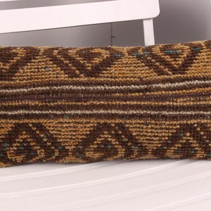 May include: Rectangular decorative pillow with a woven design in shades of brown, tan, and teal. The pillow features geometric patterns and horizontal stripes. It is placed on a white chair.
