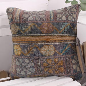 May include: Square decorative pillow with a woven, textured design. The pillow features a mix of colors including blue, brown, and gold, with geometric and floral patterns. It is placed on a white chair.