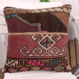May include: A decorative pillow with a geometric pattern in shades of brown, burgundy, pink, and teal. The pillow is rectangular and appears to be made of woven fabric, with a detailed design. It is placed on a white chair.