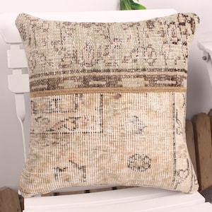 May include: A rectangular pillow with a vintage, distressed look. The top half features a patterned design in shades of beige, brown, and green. The bottom half has a lighter beige background with faded geometric patterns. The pillow is placed on a white chair.