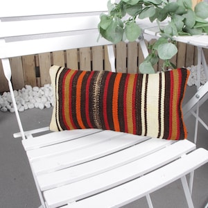 May include: Rectangular decorative pillow with a striped pattern in shades of brown, red, orange, and cream. The pillow is placed on a white folding chair. The pillow is approximately 20 inches long.