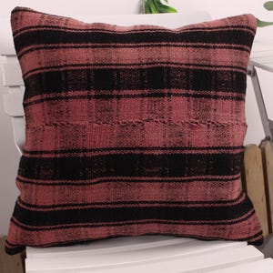 May include: A square pillow with a woven texture, featuring alternating horizontal stripes of black and a muted reddish-pink. The pillow is placed on a white chair, showcasing its decorative design and color palette.