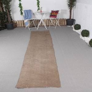 Könnte beinhalten: Ein langer, rechteckiger, hellbrauner Teppich mit gefranstem Rand. Der Teppich liegt auf einer grauen Oberfläche, mit weißen Stühlen und einem kleinen Tisch im Hintergrund. Der Teppich scheint aus einem gewebten Material zu bestehen.