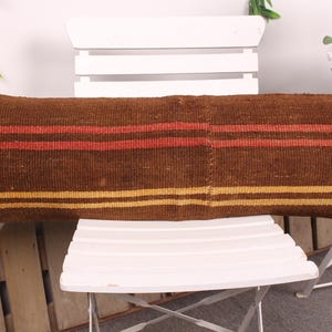 Puede incluir: Un cojín largo y rectangular marrón con rayas horizontales rojas y amarillas. El cojín está hecho de tela tejida y descansa sobre una silla blanca. Es un artículo decorativo.
